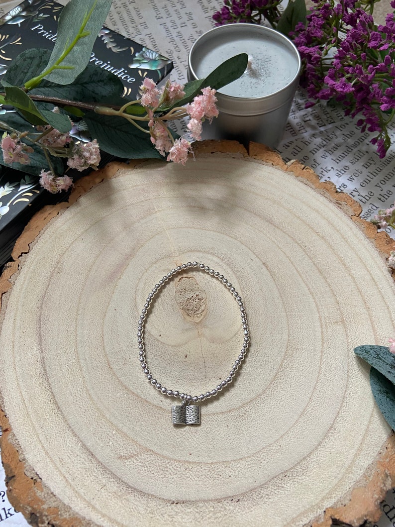 May include: A silver bracelet with a small book charm. The bracelet is on a wooden surface with a book and flowers in the background.