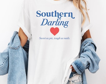 Może przedstawiać: Biała koszulka z niebieskim napisem "Southern Darling" i czerwonym sercem. Tekst pod sercem brzmi "Sweet as pie, tough as nails."