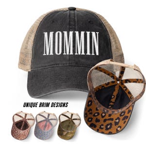 May include: A black trucker hat with a mesh back and the white text MOMMIN embroidered on the front. A second hat is shown from the underside, revealing a brown and black leopard print pattern on the brim and interior panels of the cap.