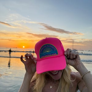 Pu&ograve; includere: Cappellino da camionista rosa acceso con la scritta "CHASING SUNSETS vacation club" su una toppa. Il cappellino ha il retro in rete e una visiera curva. L'immagine mostra il cappellino davanti a un tramonto sull'oceano.
