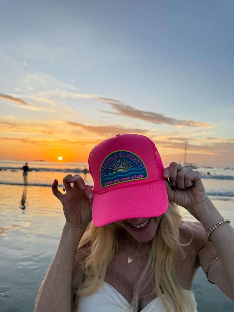 Pu&ograve; includere: Cappellino da camionista rosa acceso con la scritta "CHASING SUNSETS vacation club" su una toppa. Il cappellino ha il retro in rete e una visiera curva. L'immagine mostra il cappellino davanti a un tramonto sull'oceano.