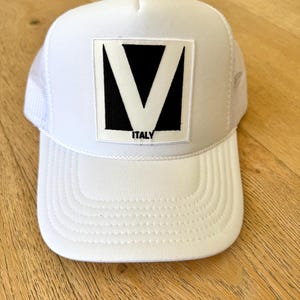 May include: White trucker hat with a black and white graphic of a V shape inside a square, with the word "ITALY" below. The hat has a white mesh back and a curved brim.