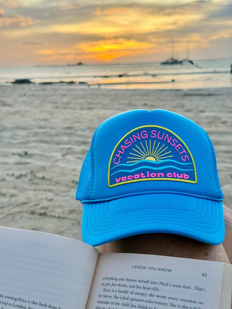 Pu&ograve; includere: Un cappellino da camionista blu brillante con la scritta "CHASING SUNSETS vacation club" in un design circolare. Il cappello ha un retro in rete e una visiera curva. Lo sfondo mostra una scena di spiaggia con un tramonto.
