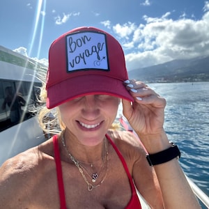 May include: A red trucker hat with a white square patch that says "Bon voyage" in black cursive writing. The hat is worn by a person with blonde hair and a red top.