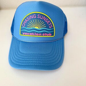 Pu&ograve; includere: Cappellino da camionista blu con visiera curva e retro in rete. Il cappello presenta una toppa con la scritta "CHASING SUNSETS vacation club" in rosa e giallo, con una grafica a forma di sole. Il cappello &egrave; di colore blu.