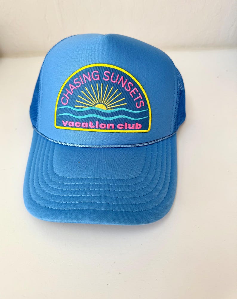 Pu&ograve; includere: Cappellino da camionista blu con visiera curva e retro in rete. Il cappello presenta una toppa con la scritta "CHASING SUNSETS vacation club" in rosa e giallo, con una grafica a forma di sole. Il cappello &egrave; di colore blu.