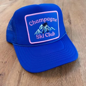 May include: A royal blue trucker hat with a mesh back and a curved brim. The front features a rectangular patch with the words "Champagne Ski Club" in pink, along with a mountain and skier graphic.