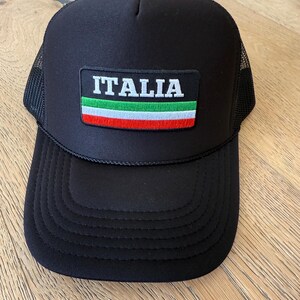 May include: Black trucker hat with a mesh back and a curved brim. The front features a rectangular patch with the word "ITALIA" in white letters above the Italian flag. The flag has green, white, and red stripes.