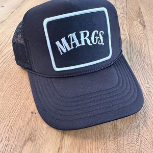 May include: Black trucker hat with a white embroidered patch that says "MARGS" in a vintage style font.