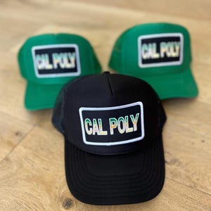 May include: Three trucker hats with a black, green, and black and green color scheme. The hats have a mesh back and a front panel with a white patch that says "CAL POLY" in rainbow lettering.