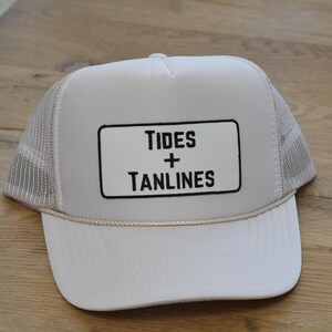 May include: A white trucker hat featuring a rectangular patch on the front panel with the black text Tides + Tanlines. The cap includes a mesh back, a solid front panel, and a thin decorative cord running across the base of the front crown.