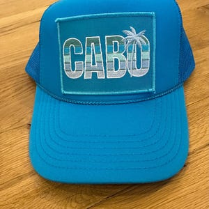 May include: A turquoise blue trucker hat with a mesh back. The front of the hat has a turquoise blue patch with the word "CABO" in white letters and a palm tree graphic.