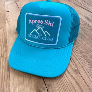May include: A turquoise trucker hat with a black mesh back. The hat has a square patch with the text "Apres Ski Social Club" in white and pink lettering. The patch features a mountain range with a cocktail glass on top.