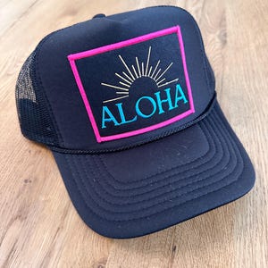 May include: Black trucker hat with a pink and blue embroidered patch that says "ALOHA" with a sun graphic.