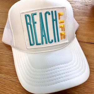 May include: White trucker hat with a white patch that says "Beach Life" in teal and gold lettering.