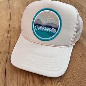 May include: A beige trucker hat with a blue and white embroidered patch that says "California" and depicts a mountain range with a sunset.