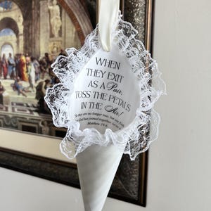 May include: A white cone-shaped petal holder featuring a ruffled lace trim and a cream-coloured ribbon loop. The front is printed with the black text: WHEN THEY EXIT AS A PAIR, TOSS THE PETALS IN THE AIR! So they are no longer two, but one flesh. God has joined together, let no one separate. Matthew 19:6.
