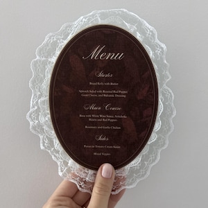 May include: An oval-shaped menu card with a dark brown background and white text, featuring sections for Starter, Main Course, and Sides. The card is framed by a delicate white lace border that extends beyond the edges of the oval paper.