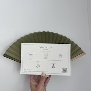 Könnte beinhalten: Eine rechteckige weiße Hochzeits-Zeitplan-Karte für Jacob & Larissa mit schwarzem Text, Veranstaltungssymbolen und einem QR-Code, präsentiert vor einem gefalteten olivgrünen Fächer. Die Karte listet Ereignisse von 18:00 bis 22:00 Uhr auf, einschließlich Zeremonie und Empfang.