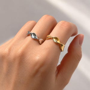 Peut inclure: Deux bagues au design ondulé et torsadé. Une bague est de couleur argentée et portée sur l'index, tandis que l'autre est de couleur dorée et portée sur le majeur. Les deux anneaux présentent une surface lisse et réfléchissante aux courbes irrégulières.