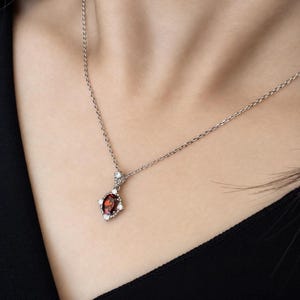 May include: A silver-toned necklace featuring a delicate chain and a pendant with a central oval-cut deep red gemstone. The stone is surrounded by four small, clear, round-cut accent stones set in a metallic frame with a pointed design.