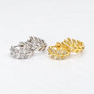 May include: A pair of silver-toned hoop earrings and a pair of gold-toned hoop earrings, both featuring a leaf-shaped design encrusted with small clear stones. The silver earrings are stamped with S925, indicating sterling silver construction.