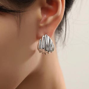 May include: Silver tone earrings with a geometric design. The earrings are shaped like a small, rounded triangle with multiple horizontal lines.