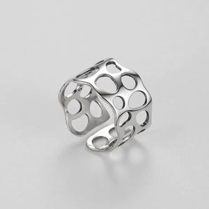 May include: A silver ring with a unique, openwork design featuring a repeating pattern of circles. The ring is made of a smooth, polished metal and has a modern, minimalist aesthetic.
