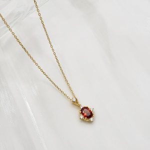 May include: A gold chain necklace with a pendant featuring a red oval gemstone surrounded by smaller clear gemstones.