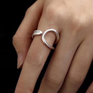 Peut inclure: Une bague argentée au design ouvert unique. La bague présente une forme incurvée et fluide qui s'enroule autour du doigt. La bague est faite d'un matériau métallique brillant et est présentée sur une main.