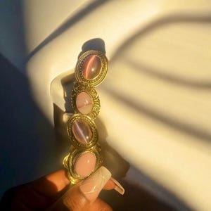 May include: Gold hair clip with four oval-shaped iridescent stones. The stones are pink, purple, and gray.