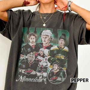 Könnte beinhalten: Dunkelgraues T-Shirt mit einer Grafik von Eishockeyspielern, dem Wort "WILD" in Grün und "Minnesota" in Weiß. Das Shirt hat einen Vintage-Look.