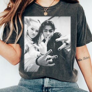 May include: Dark grey t-shirt with a black and white photograph of two people making hand gestures. The photo is rectangular and centred on the shirt. The shirt has a crew neck and short sleeves. The person wearing the shirt is also wearing blue jeans.