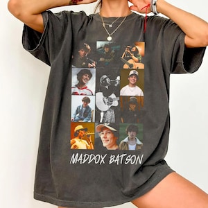Puede incluir: Camiseta gris oscuro con una cuadrícula de nueve imágenes de una persona, con el nombre "MADDOX BATSON" impreso en blanco debajo. Las imágenes muestran a la persona en varias poses, algunas tocando instrumentos.