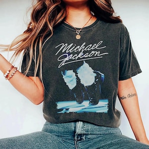 May include: Dark gray t-shirt featuring the name "Michael Jackson" in white script above a graphic of the singer's silhouette. The shirt has a vintage, worn look. The model is wearing blue jeans.