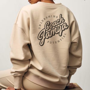 May include: A beige sweatshirt with a circular graphic on the back. The graphic features the text "Coach Jamya" in a stylized font, surrounded by the text "Unleashing Potential".