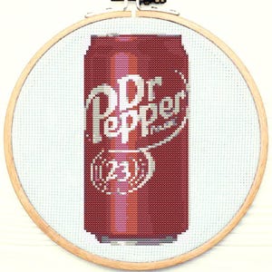 May include: A cross-stitch embroidery of a red Dr Pepper can with the brand name and 23 written on it. The embroidery is framed in a wooden hoop.