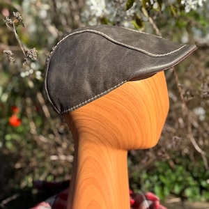 May include: A brown leather flat cap with a contrasting white stitch detail. The cap is on a wooden mannequin head.