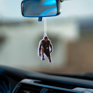 May include: A car air freshener featuring a basketball player in a black and white jersey with the number "3" on it. The air freshener is hanging from the rearview mirror of a car.