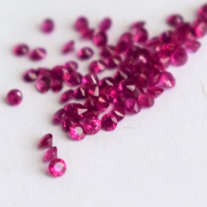 Pode incluir: Uma dispersão de pequenas pedras preciosas de rubi redondas e facetadas em vários tamanhos. As pedras são de uma cor vermelha profunda e vibrante e estão espalhadas sobre uma superfície branca, criando um efeito brilhante. As pedras preciosas são provavelmente para fazer joias.