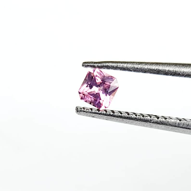 May include: A small, square-cut pink gemstone with multiple facets that reflect light, held securely between the two serrated metal tips of a pair of silver-coloured tweezers against a plain, bright white background.