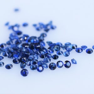 Op de afbeelding: Een verzameling kleine, ronde, gefacetteerde blauwe saffieren verspreid over een effen wit oppervlak. Elke edelsteen vertoont een diepe, levendige blauwe tint met meerdere facetten die het licht weerkaatsen, wat zorgt voor een sprankelend effect.