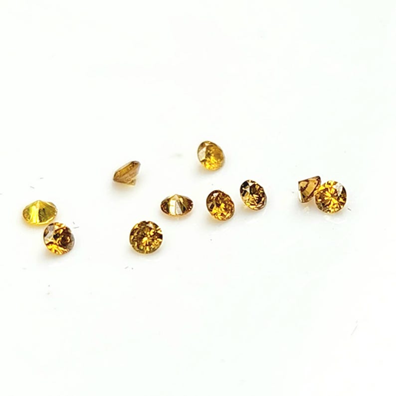 May include: A collection of nine small, round, faceted gemstones in shades of golden yellow and amber. Each stone features a brilliant cut with multiple reflective surfaces that catch the light, creating a sparkling appearance against a plain white background.