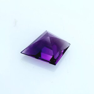 May include: A square-cut, deep purple gemstone with a faceted pyramid shape. The stone features a pointed apex and flat, angled sides that create a geometric silhouette. The translucent material displays varying shades of violet and dark purple.