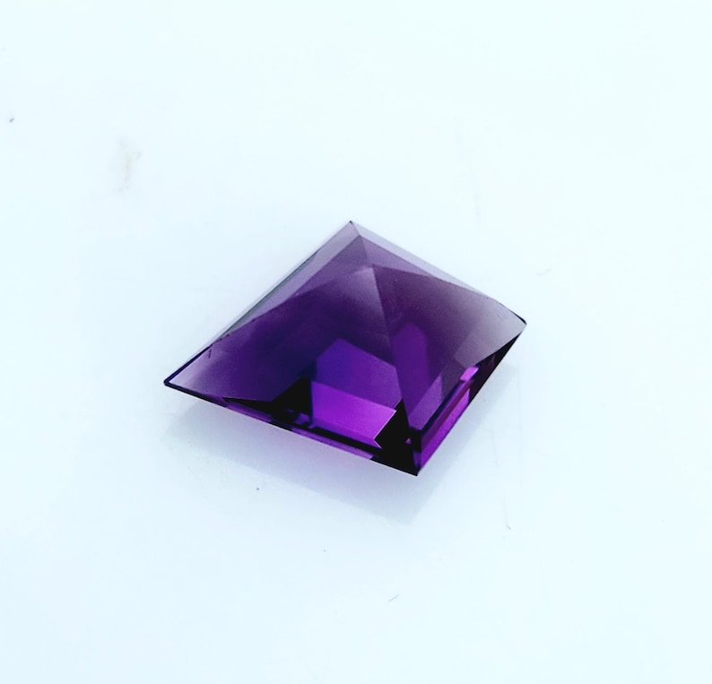 May include: A square-cut, deep purple gemstone with a faceted pyramid shape. The stone features a pointed apex and flat, angled sides that create a geometric silhouette. The translucent material displays varying shades of violet and dark purple.