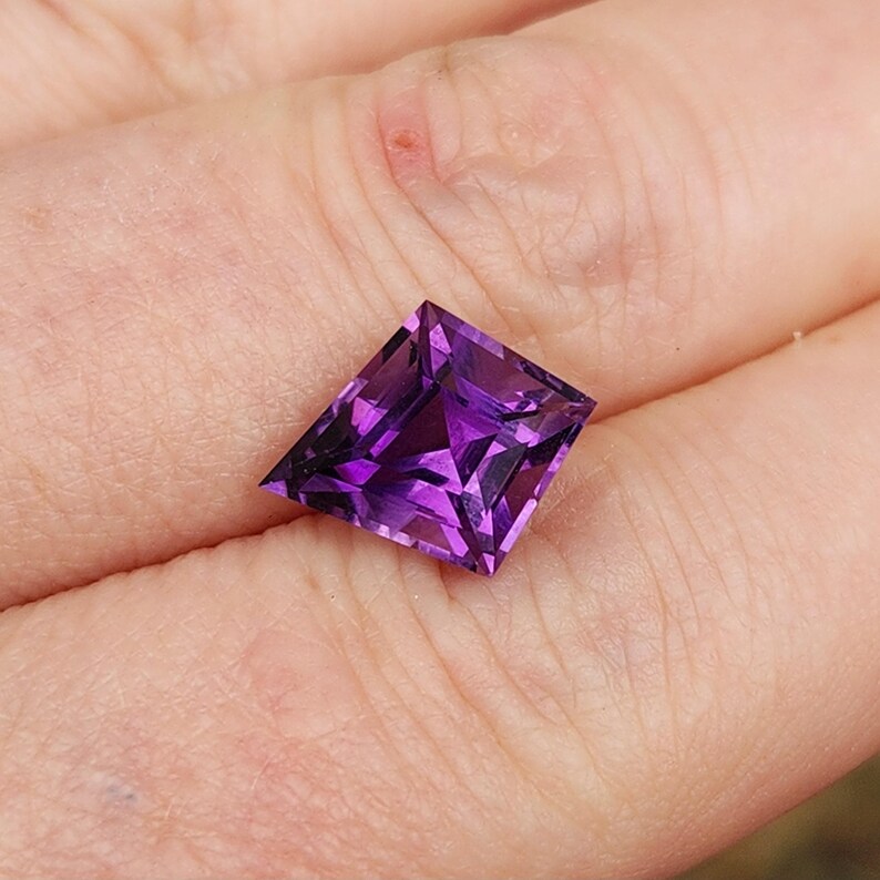 May include: A kite-shaped, faceted gemstone with a deep, vibrant purple color. The stone features a symmetrical geometric cut with sharp, pointed corners and a brilliant, reflective surface that displays various shades of violet and dark purple.