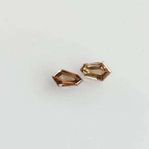 May include: Two loose shield-cut gemstones with a warm, light brown hue. Each stone features a faceted surface that reflects light, displaying a geometric shape with a pointed end and a flat top edge, showcasing a translucent, amber-like clarity.