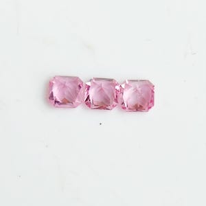 May include: Three square-cut, translucent pink gemstones arranged in a horizontal row. Each stone features a faceted surface that reflects light, creating a sparkling appearance with distinct geometric edges and a uniform light pink colour throughout.