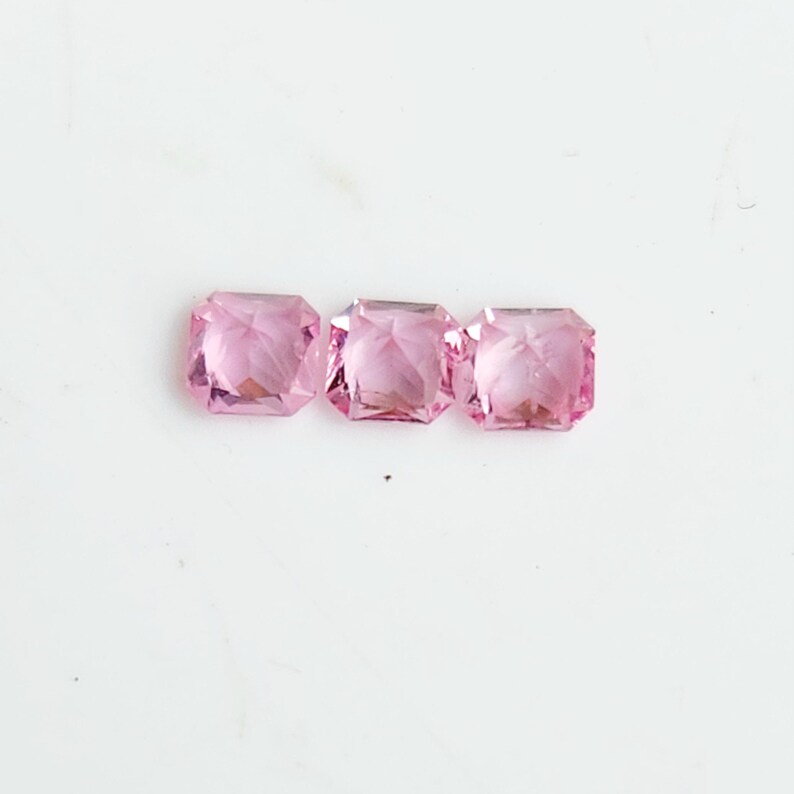 May include: Three square-cut, translucent pink gemstones arranged in a horizontal row. Each stone features a faceted surface that reflects light, creating a sparkling appearance with distinct geometric edges and a uniform light pink colour throughout.