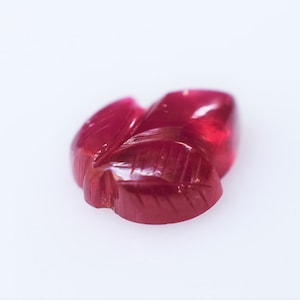 May include: A carved ruby gemstone in the shape of a leaf. The ruby is a deep red color and has a glossy finish.